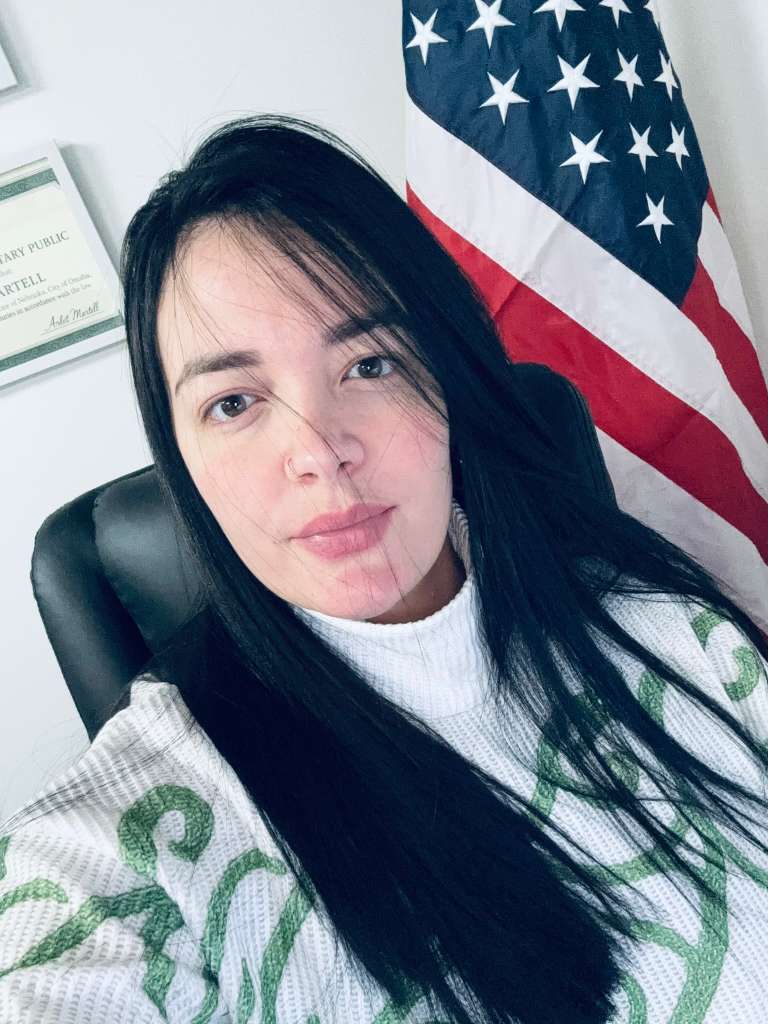 Mujer de cabello negro largo sentada frente a una bandera estadounidense y un certificado enmarcado.