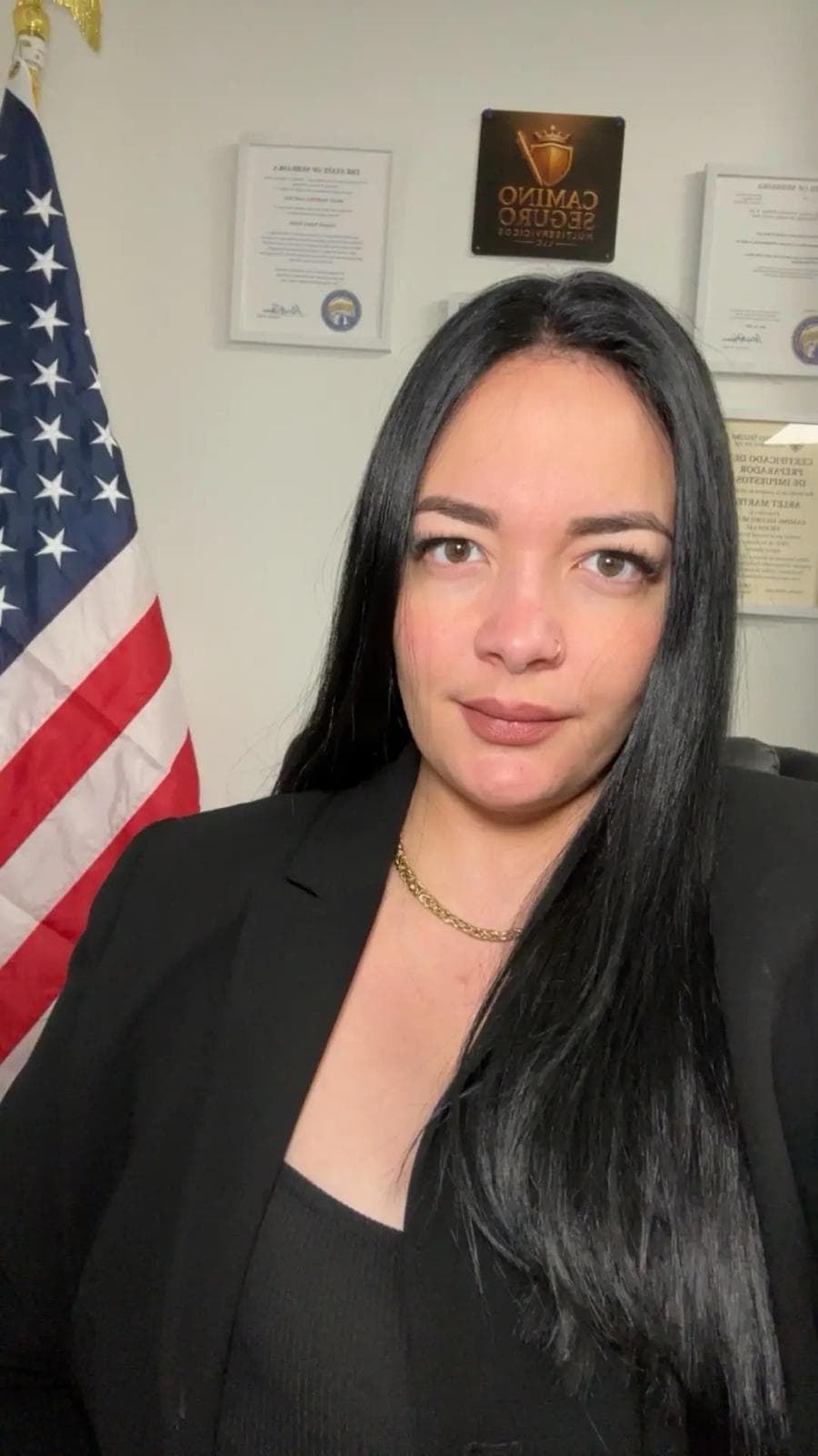 Mujer con cabello negro y chaqueta negra frente a una bandera estadounidense y diplomas.